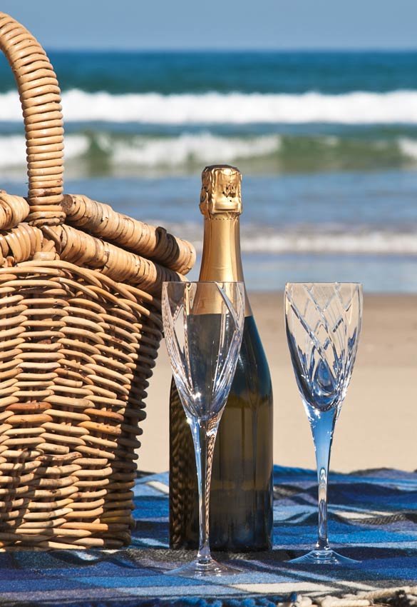 A picnic basket, a bottle of champagne, and two empty crystal glasses sit on a blanket on the beach, a relaxing addition to things to do in Port St. Joe, Florida.