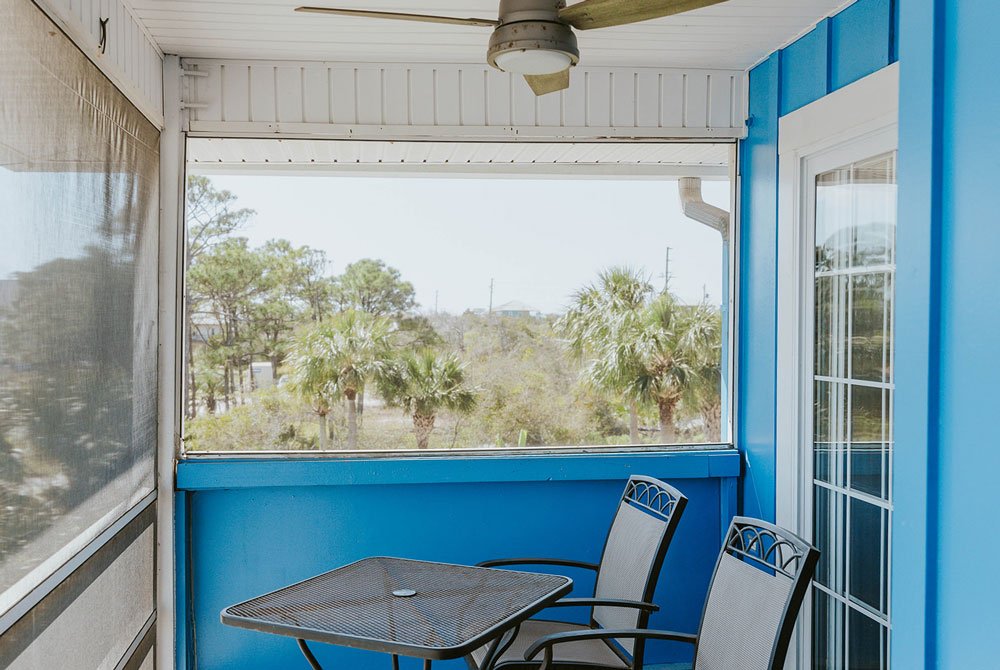 A screened-in porch with bright blue walls at the Turtle Shell Suite, featuring a metal patio table and two chairs overlooking a wooded landscape.