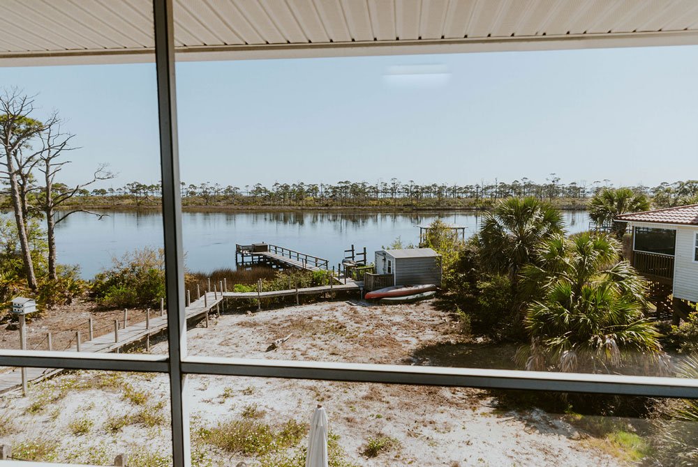 View from the Turtle Shell Suite porch overlooking a sandy yard, a small dock on a calm river, and a line of pine trees in the distance.