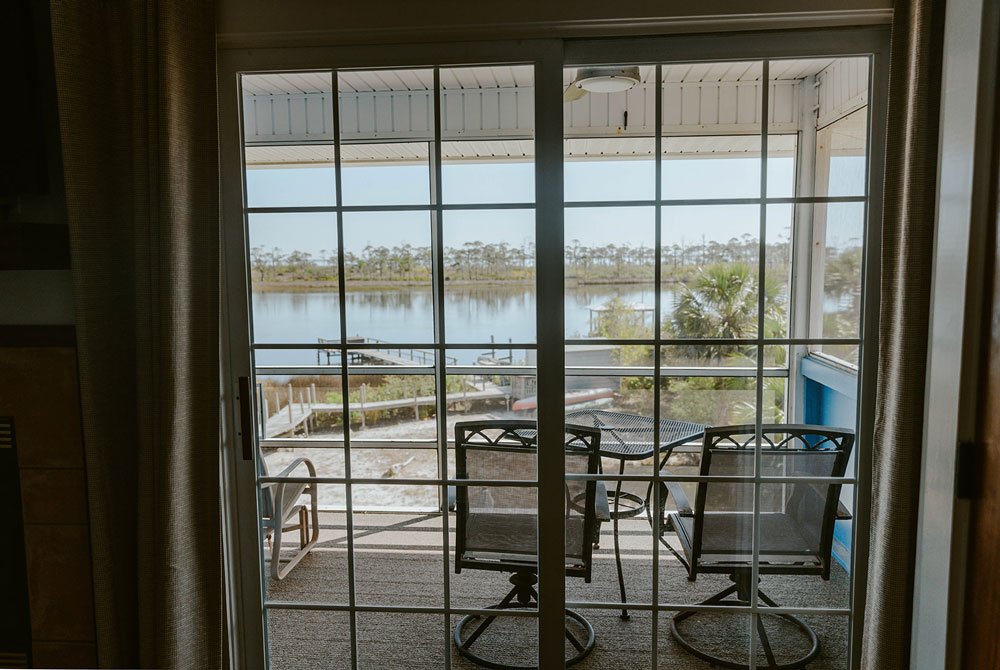 A view through sliding glass doors of the Turtle Shell Suite porch, featuring outdoor chairs overlooking a lake and a wooden dock.