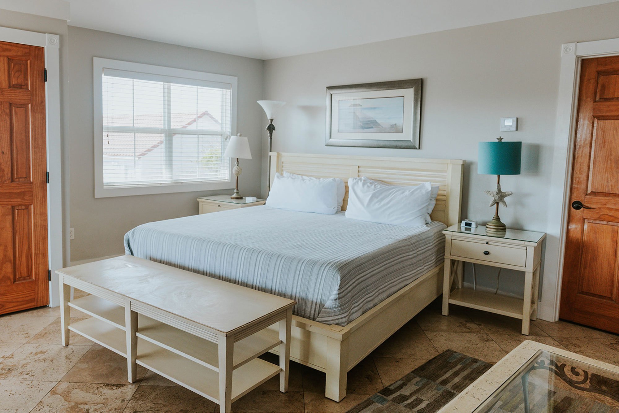 A bedroom at the Turtle Shell Suite Cape San Blas Inn with a light-colored wood bed frame, striped bedding, two matching nightstands with lamps, and a wooden bench at the foot of the bed.