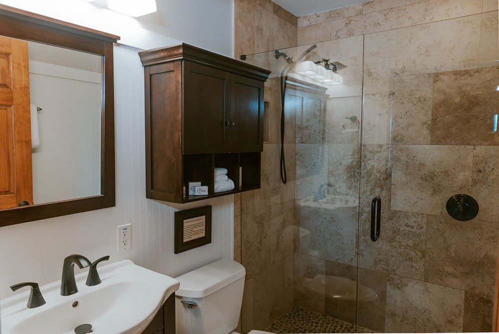 A bathroom vanity with a dark wood cabinet, white sink, and glass-enclosed walk-in shower with stone tile walls at the Turtle Shell Suite in the Cape San Blas Inn.
