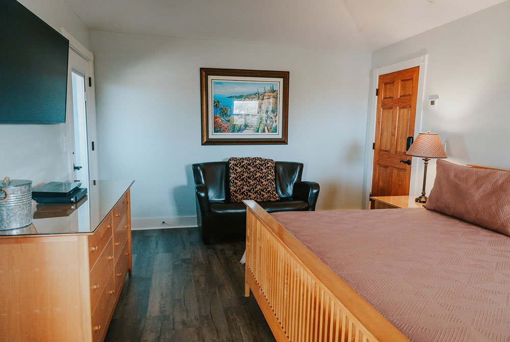 Bedroom at the Cape San Blas Inn featuring a bed with a light wood frame, a dark leather loveseat, a light wood dresser, and a framed painting on the wall.