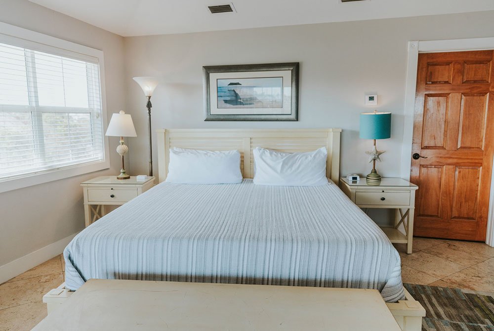 A Turtle Shell Suite bedroom at the Cape San Blas Inn featuring a king bed with striped bedding, two nightstands with lamps, a framed coastal print above the headboard, and a wooden door.