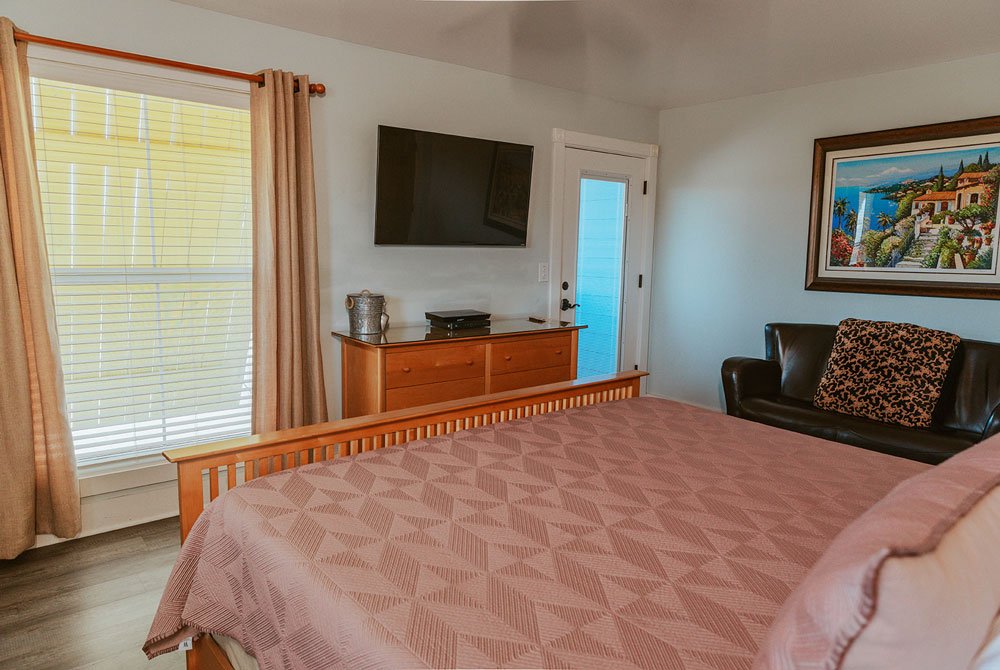 A bedroom at the Cape San Blas Inn featuring a bed with a geometric-patterned pink quilt, a wooden dresser with a wall-mounted television, a leather armchair with a patterned pillow, and a framed landscape painting on the wall.