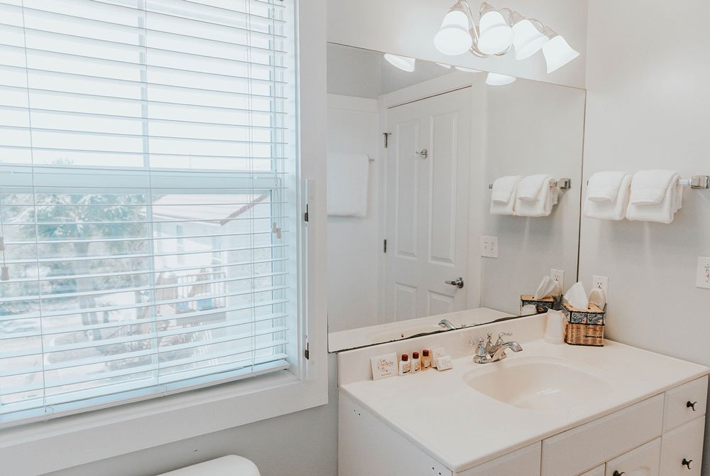 A white bathroom vanity with a sink, mirror, and toiletries at the Cape San Blas Inn, situated next to a window with blinds.