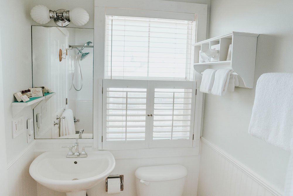 A bright, all-white bathroom at the Coquina Shell Suite, featuring a pedestal sink, a toilet, a window with plantation shutters, and a wall-mounted storage shelf with folded towels.