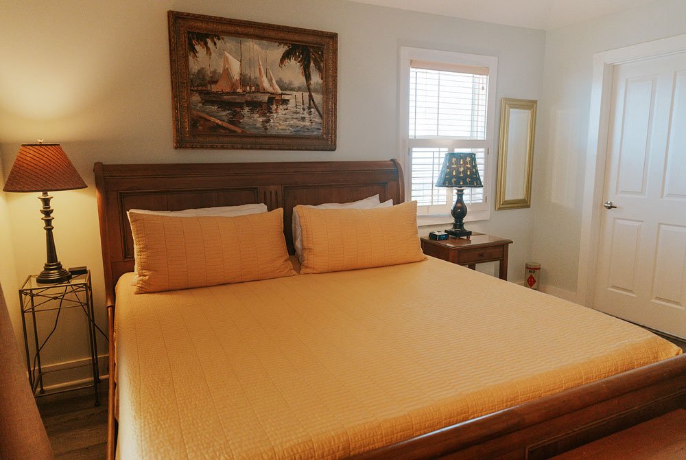 A bedroom in the Coquina Shell suite features a king-sized bed with a yellow quilt, a framed nautical painting above the headboard, and two bedside lamps.