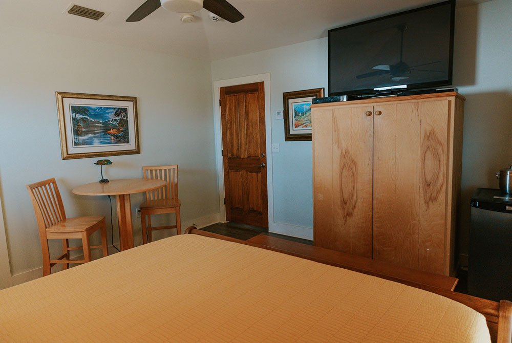 A bedroom in the Coquina Shell Suite featuring a bed with a tan quilt, a small wooden dining table with two chairs, a tall wooden wardrobe topped with a television, and a wooden door.
