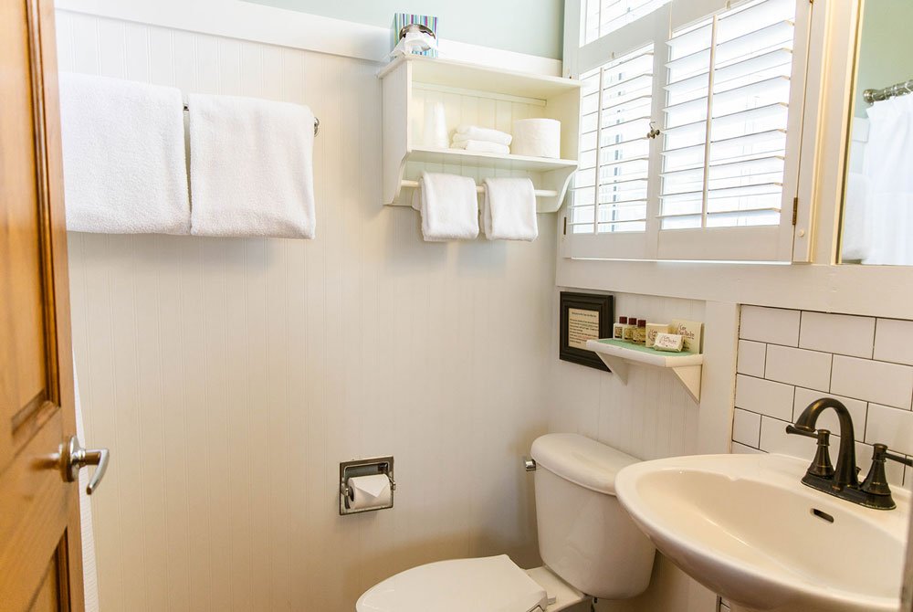 A small bathroom at the Scallop Shell room featuring a toilet, a pedestal sink with a dark faucet, white wall-mounted shelving with towels and toiletries, and white shutters over a window.