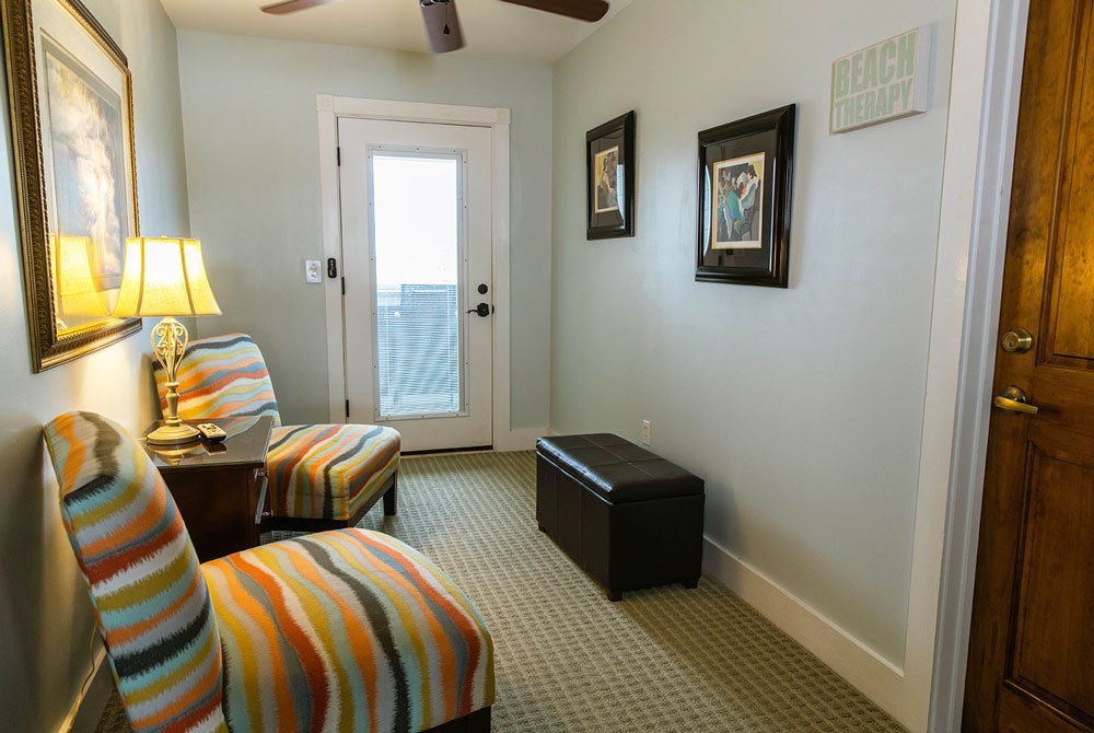 A hotel room seating area in the Coquina Shell Suite with two striped armchairs, a small side table with a lamp, a leather storage ottoman, and a sign on the wall that reads “Beach Therapy.”