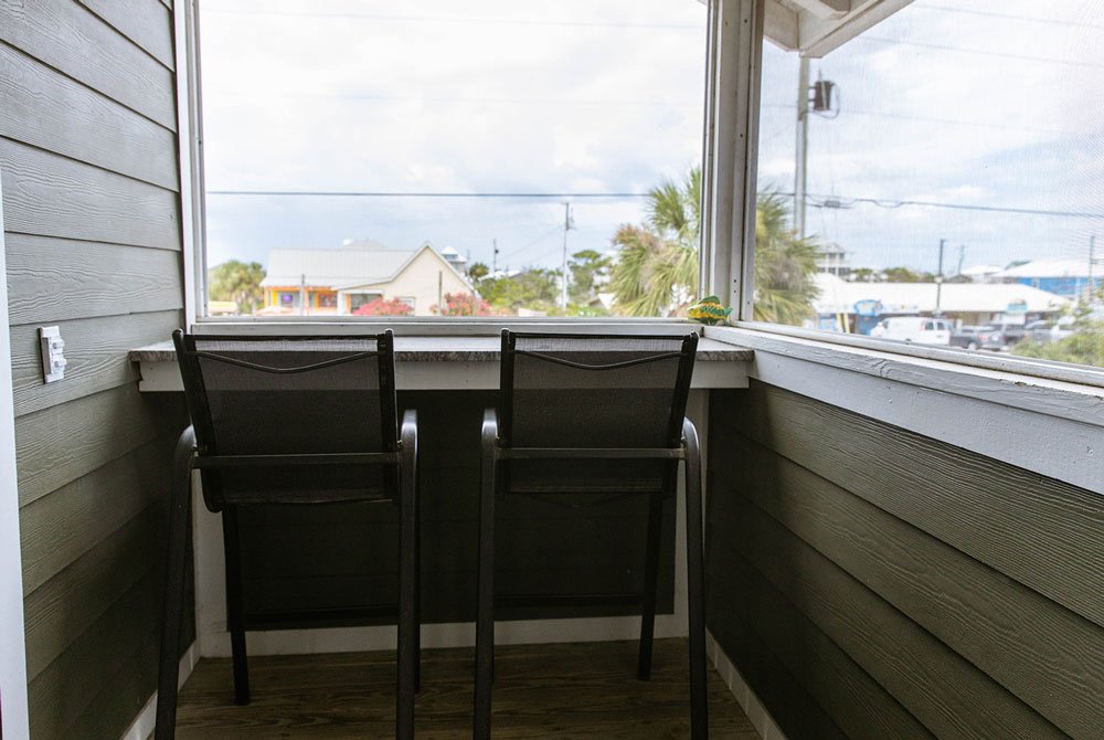Two chairs face a window counter on the screened-in porch of the Coquina Shell Suite, looking out at a street with houses and palm trees.