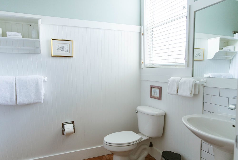 A bright white bathroom with beadboard walls, a toilet, a pedestal sink, and a wall-mounted towel rack in an oyster shell room at the Cape San Blas Inn.
