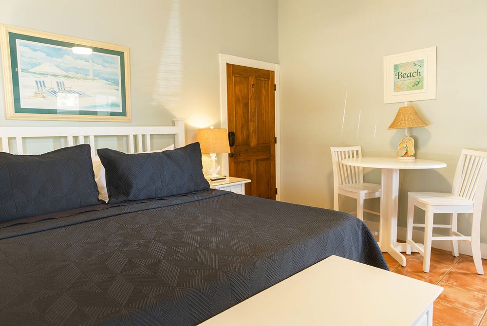 The Sand Dollar room at the Cape San Blas Inn features a queen bed with a dark navy quilted cover, a small white bistro table with two chairs, and framed wall art.