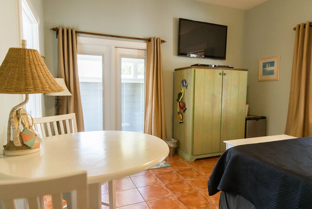 A white round table with a coastal-themed lamp, a light green wooden cabinet topped with a television, and a portion of a bed in a Cape San Blas Inn room with a glass door and beige curtains.