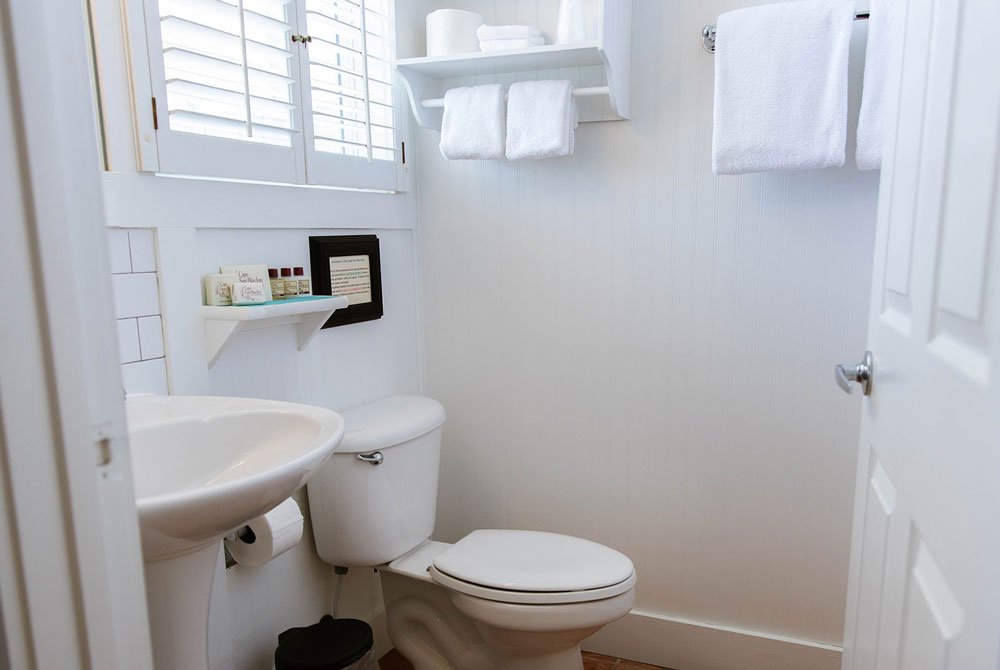 A white bathroom at the Sand Dollar Room, featuring a pedestal sink, a toilet, a wall-mounted shelf with toiletries, and towel racks with white towels.