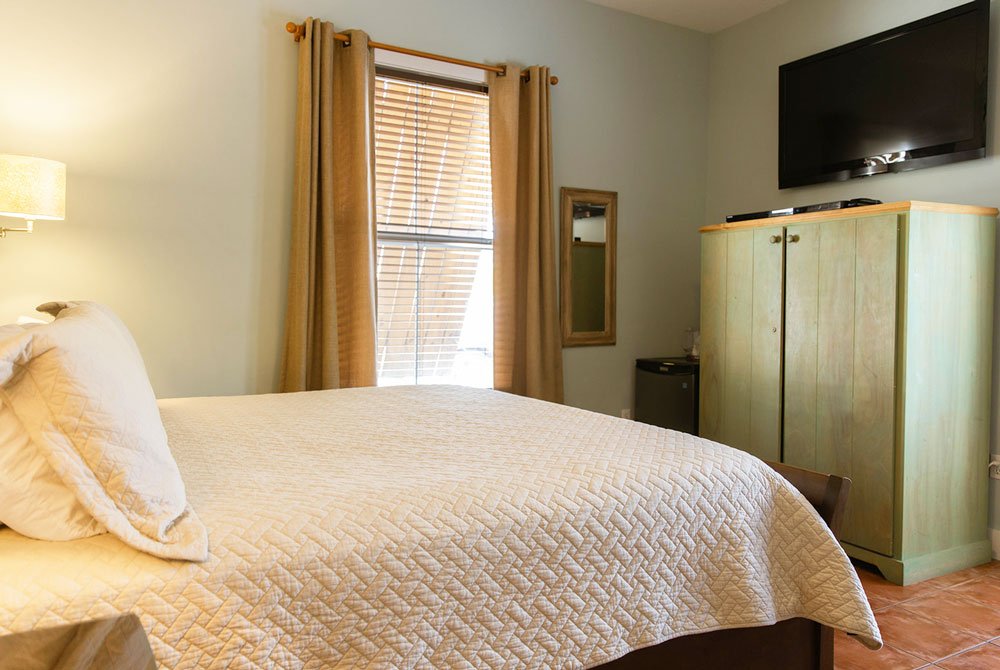 A bedroom at the Cape San Blas Inn featuring a bed with a quilted white coverlet, a large wooden wardrobe topped with a television, and a window with tan curtains.